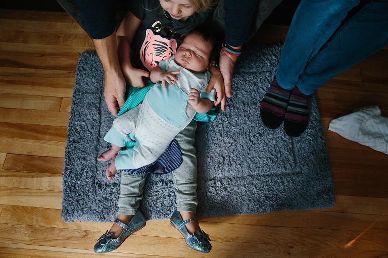 Top down view of whole family. Baby in sister's lap, Dad offers hands of support. Mom's feet at in image. 