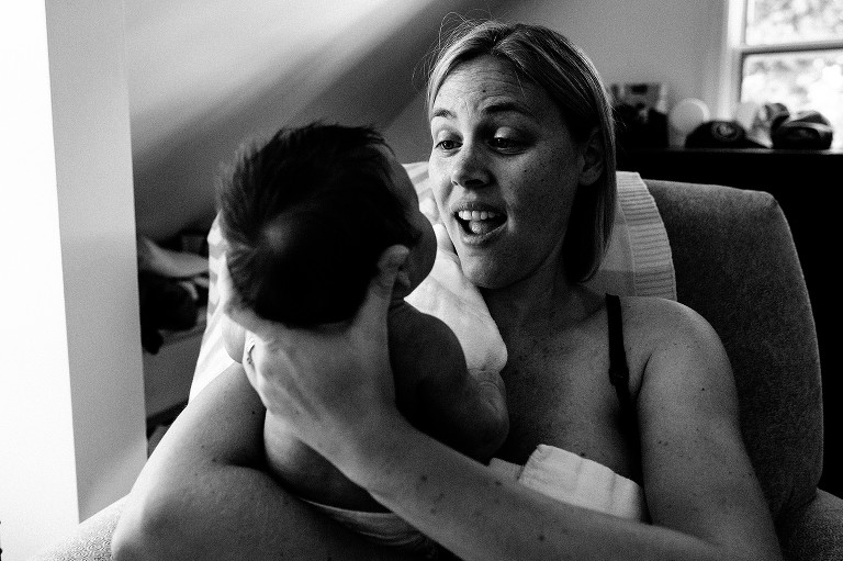 Mom smiles at newborn, sitting in the chair with subtle window light coming in camera left.