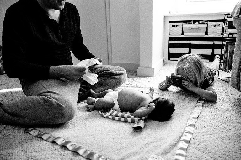 Sister takes newborn brother's picture wiht toy camera while dad starts to change his diaper 