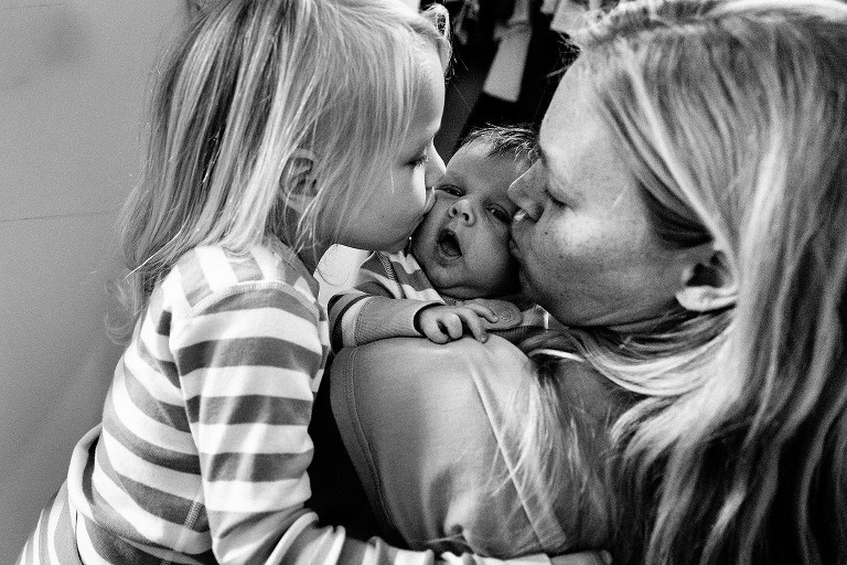 Black and white. Preschool sister and mom both kiss the cheek of new baby at hte same time. 