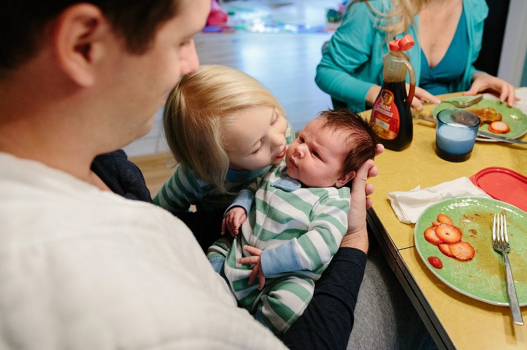 Sister comes into kiss newborn brother, baby looks at dad, mom eats breakfast 