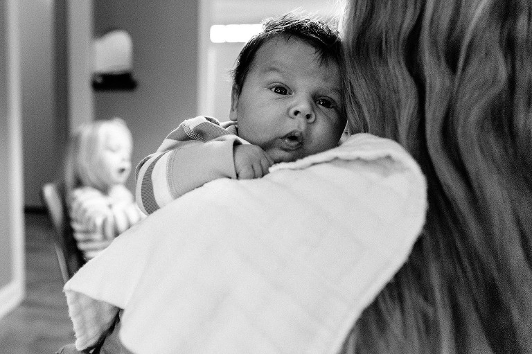 Mom holds baby over her shoulder, baby is awake, daughter in background is unknowingly making a similar mouth expression as baby brother 