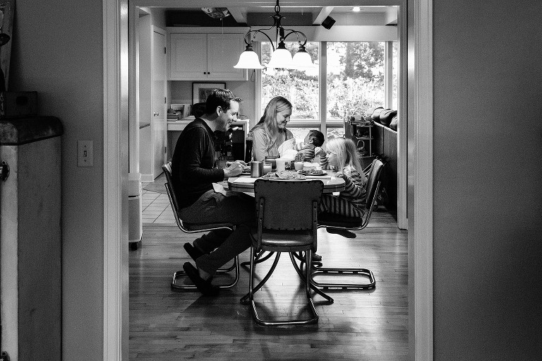 Black and white photo of family at breakfast table. Dad smiling at preschool daughter, mom smiles at newborn smile 