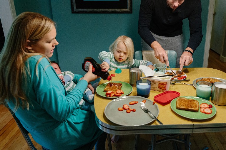 Family of four at breakfast table. Mom and preschool daughter pour syrup on mom's toast, and dad cuts daughter's French toast. 