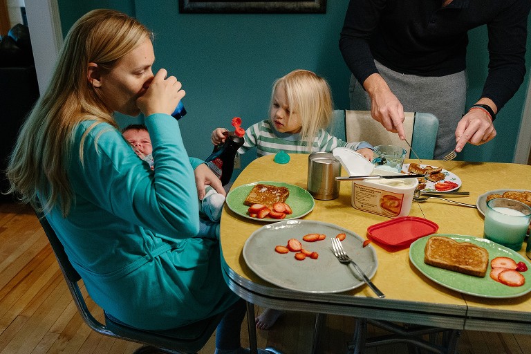 Family of four at breakfast table. Mom drinks coffee and holds newborn, preschool daughter pours syrup on mom's toast, and dad cuts daughter's French toast. 