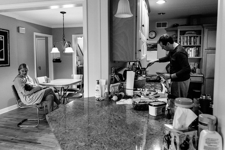 Wide scene: Mother holds newborn baby in dining room, dad flips French toast in kitchen 