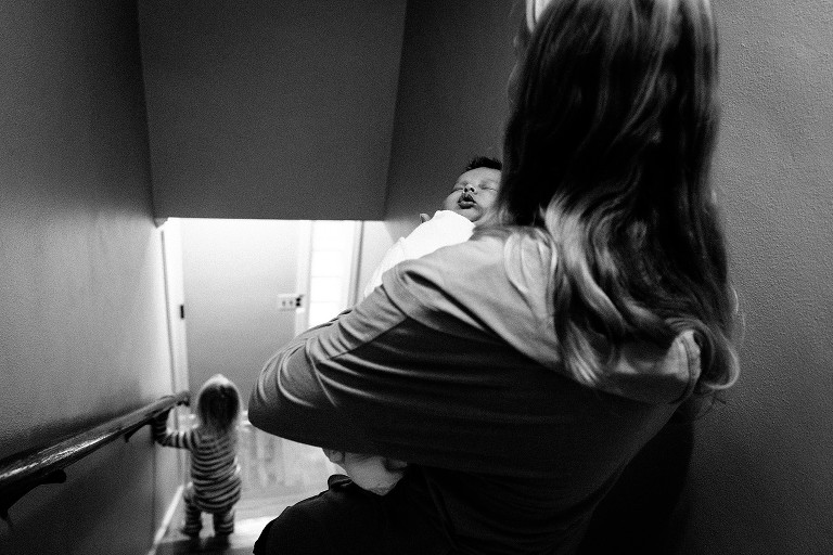Mom carries newborn downstairs while toddler is ahead of them 