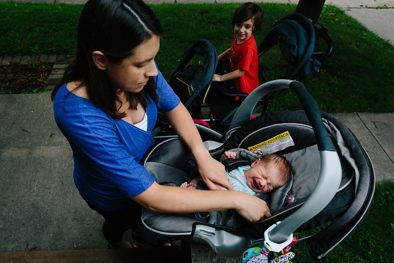 Mom straps crying newborn into carseat. Boy looks on from his standing stroller. 