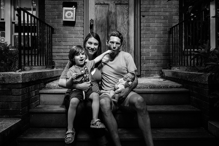 Family portrait on front steps of the house. Preschool son pretends to superhero punch his dad and dad playfully responds. 