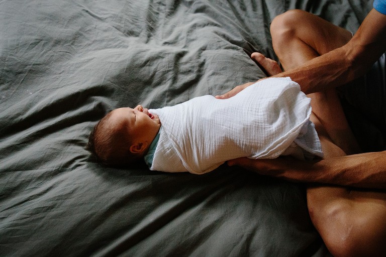 Dad swaddles newborn in white blanket on bed. 