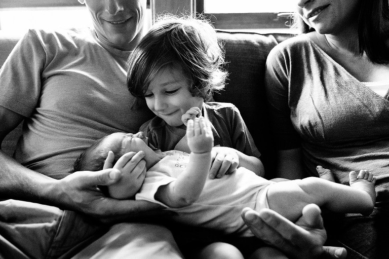 Black and white. Family on couch, preschool boy holding crying newborn, parents look on. 