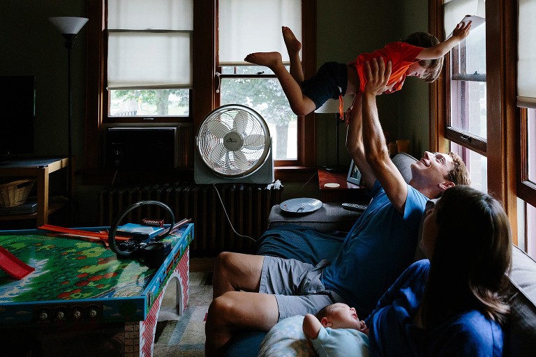 Dad tosses preschool son up above his head ot play airplane. Mom and baby in foreground. 
