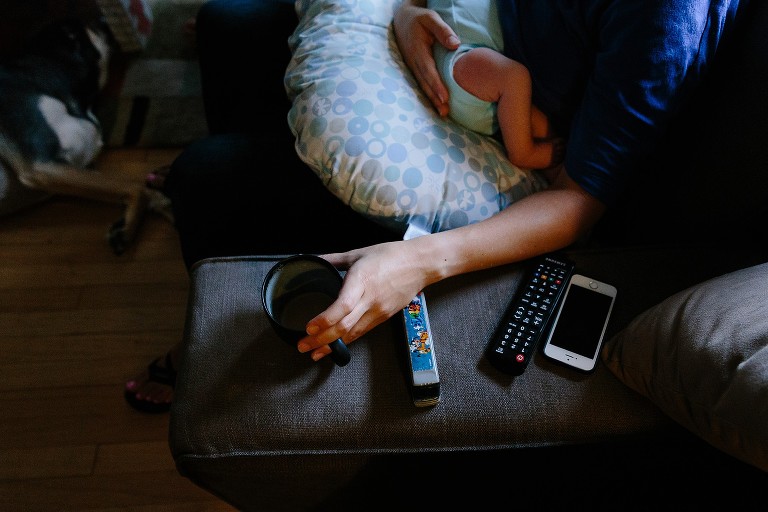 Mom holds baby while reaching for coffee. Remotes and iPhone sit on the arm rest of couch. 