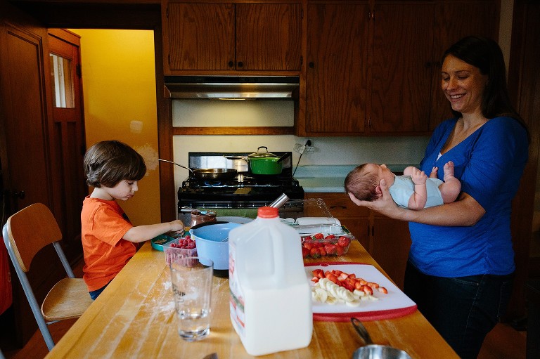 Preschool boy eats breakfast while Mom stands and holds baby, smiling. 