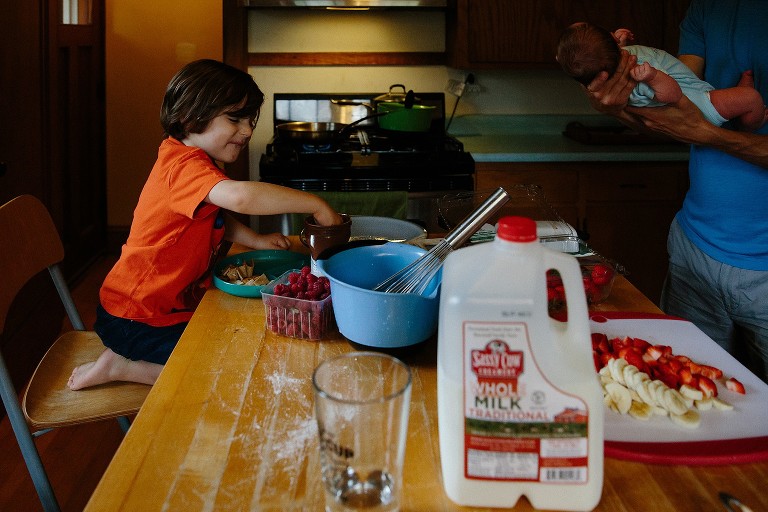 Preschool boy sticks hand in Nutella while Dad holds newborn baby across the table. 