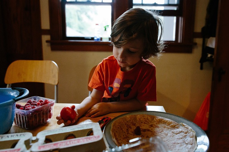 Preschool boy holds a large strawberry with large bite out. Crepes in foreground. 