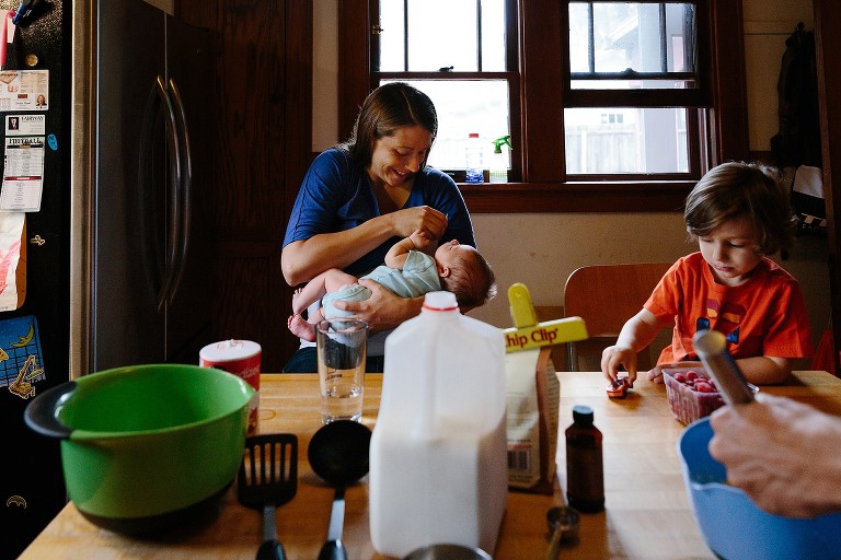 Mom engaged with nursing newborn while boy runs a toy car over the table. Dad's hand in foreground whisking batter for crepes. 