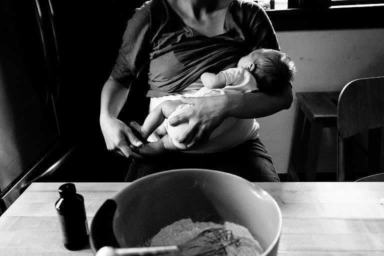 Black and white photo of mom nursing baby, holding her feet. Bowl of flour in foreground. 