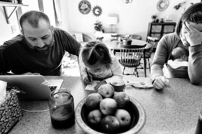 dad works on computer, toddler writes on notepad, mom holds head with a migraine while baby wearing newborn in madison newborn photo session