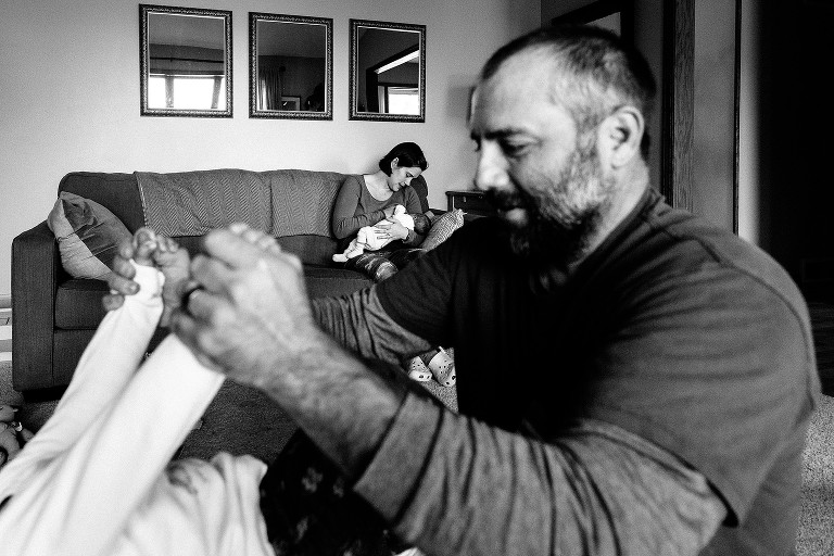 mom nurses baby on couch in background, dad plays with older sibling in foreground. Black and white