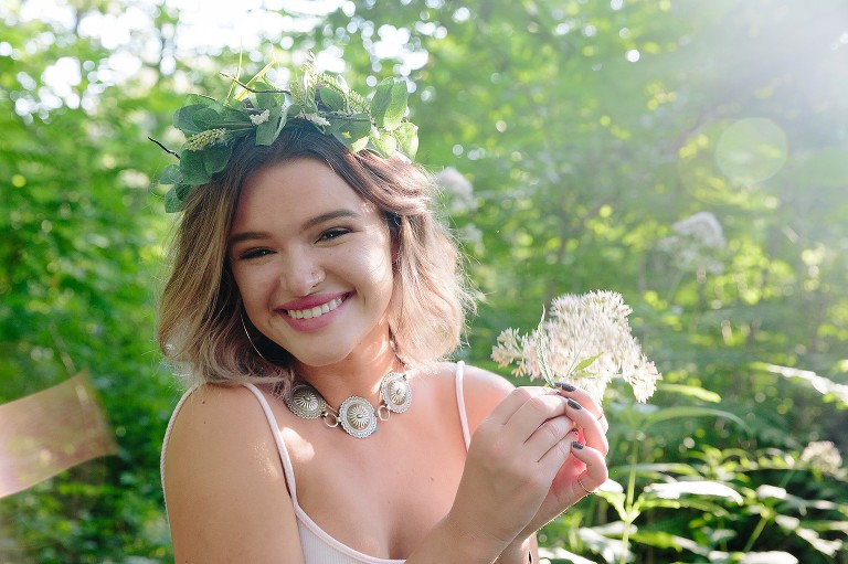Young woman smiles in forest holding flowers. Sunlight catching camera lens.