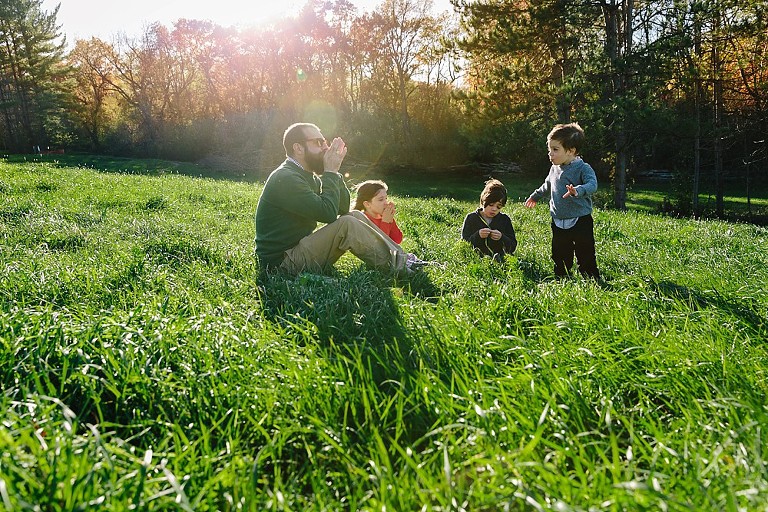 Dad demonstrates how to whistle through a blade of grass. Two older kids are eager to practice, while younger son is content to be an awed listener. 