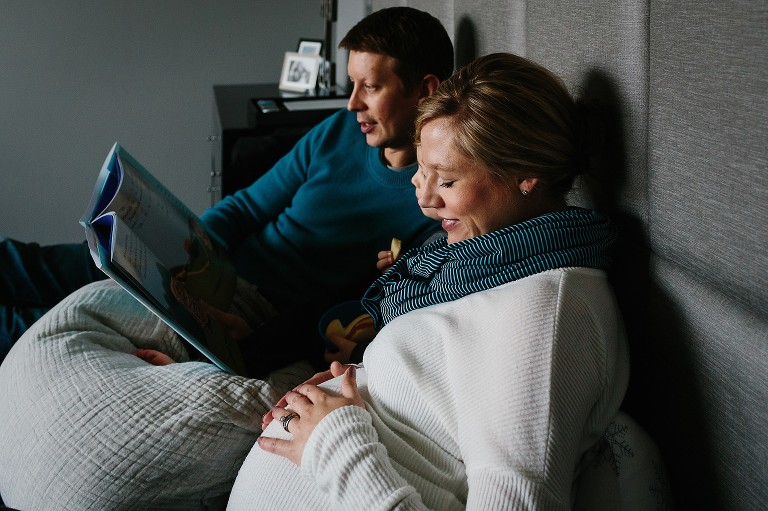 Family sits together in bed. Father reads book to son, mother looking at her baby bump.