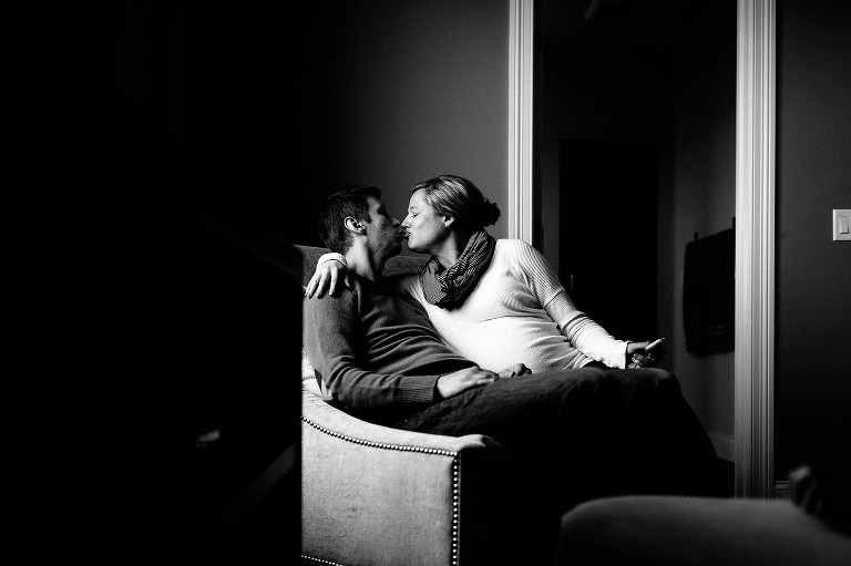 Black and white. Husband and pregnant wife kiss sitting in chair together.