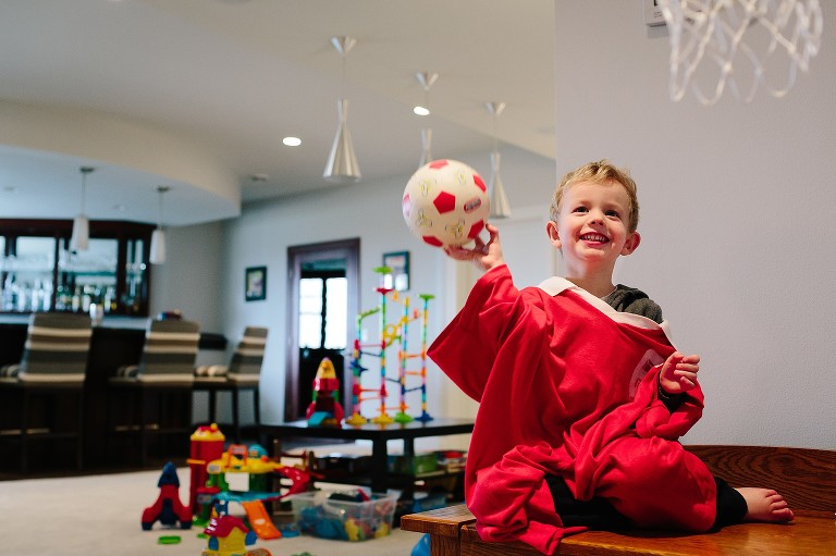 Young boy wearing fathers old soccer jerseys smiles and holds up soccer ball in his playroom.