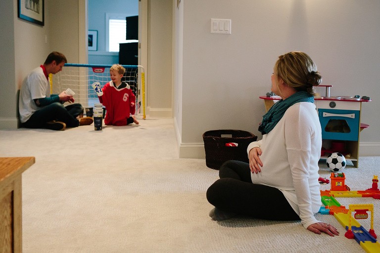 Pregnant mother watches father and son play dress up in fathers old soccer jerseys in the playroom.