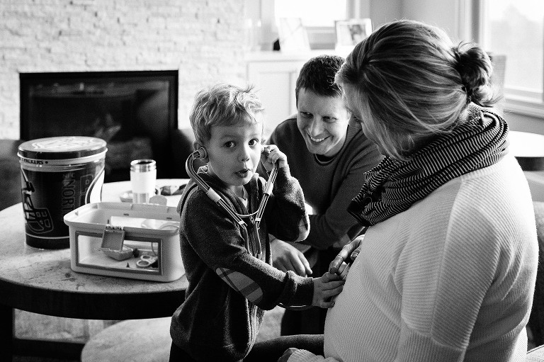 Black and white. Son plays doctor listening to pregnant moms belly, father smiles.