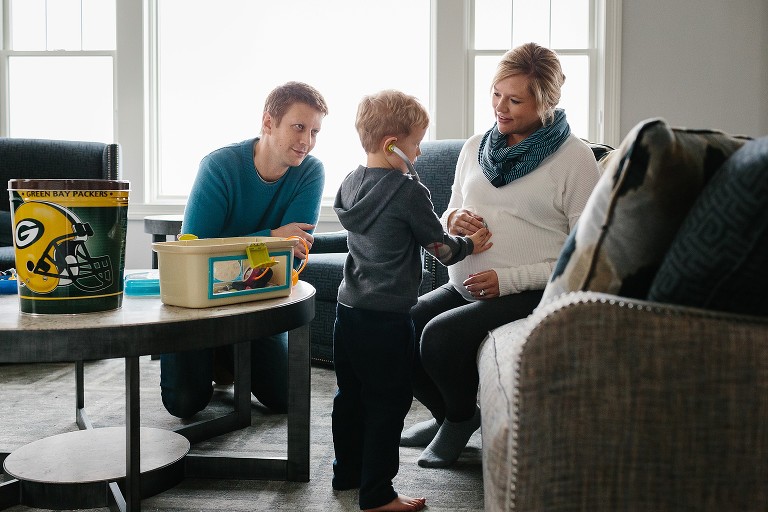 Family sits together in living room. Son plays doctor listening to pregnant mothers belly, father watching.
