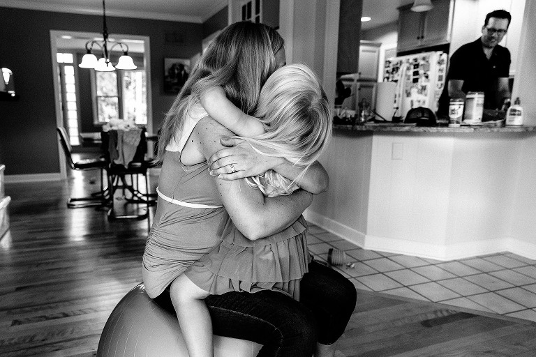 Mom sits on exercise ball and hugs daughter while dad in the background in kitchen, smiling. 