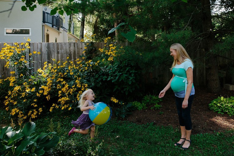Preschool girl runs toward pregnant mother in backyard. Black-eyed Susan flowers behind them. 