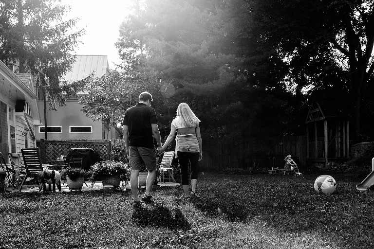 black and white. Mom and Dad walk hand in hand in backyard. Preschool girl pushes toy lawn mower in background. Summer evening sun flare present 