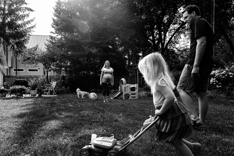 Layered scene. Girl pushes toy lawn mower, Dad, pregnant mom, and family dog in background. 