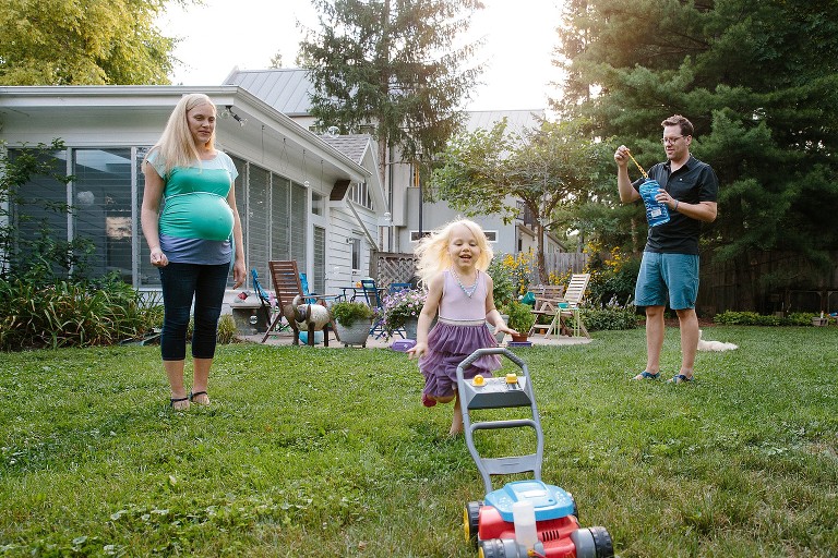 Dad blows bubbles, preschool girl chases bubble lawn mower, pregnant mom watches in backyard 
