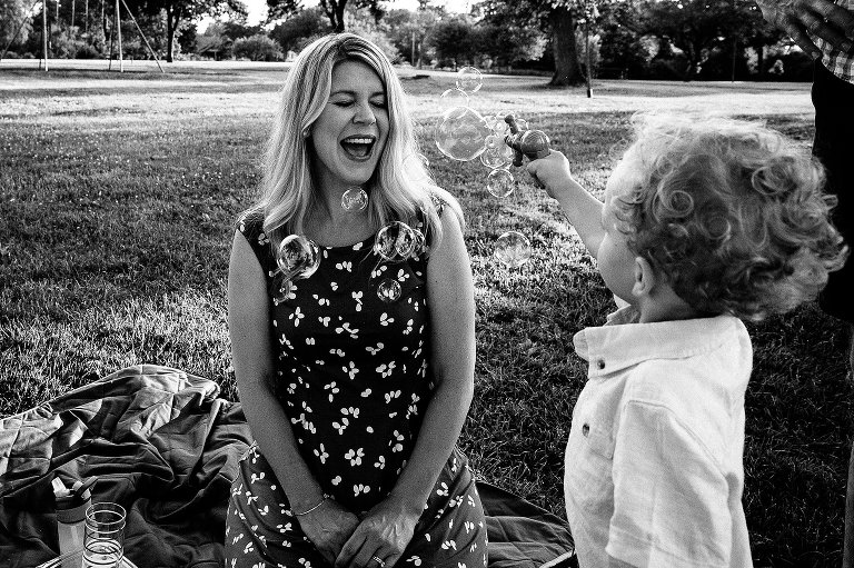 Mother closes her eyes and smiles as her son uses a bubble "gun" to shoot lots of bubbles in her direction. Black and white. 