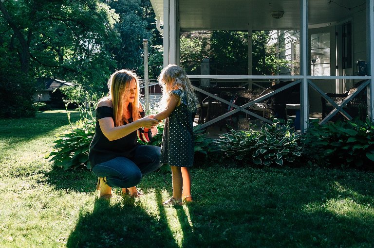 Mother helps young daughter fit her hand into her pink baseball glove. They are standing in warm, dappled afternoon light. 