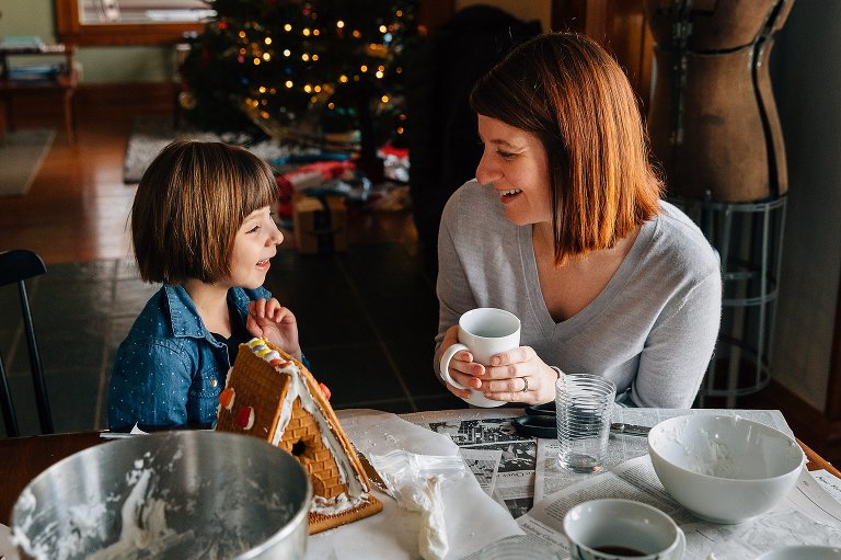 Mother holds a cup of coffee as she talks to her young daughter over a building a gingerbread house. There is a Christmas tree in the background and an A-frame ginggerbread house in the foreground. 
