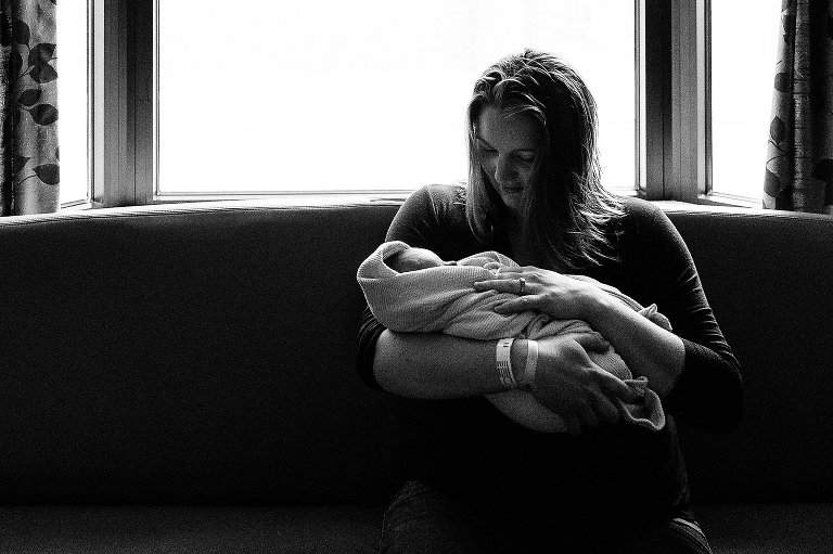Mother holds her newborn at the hospital. She is on a couch with a lot of black, negative space to the left. Black and white.