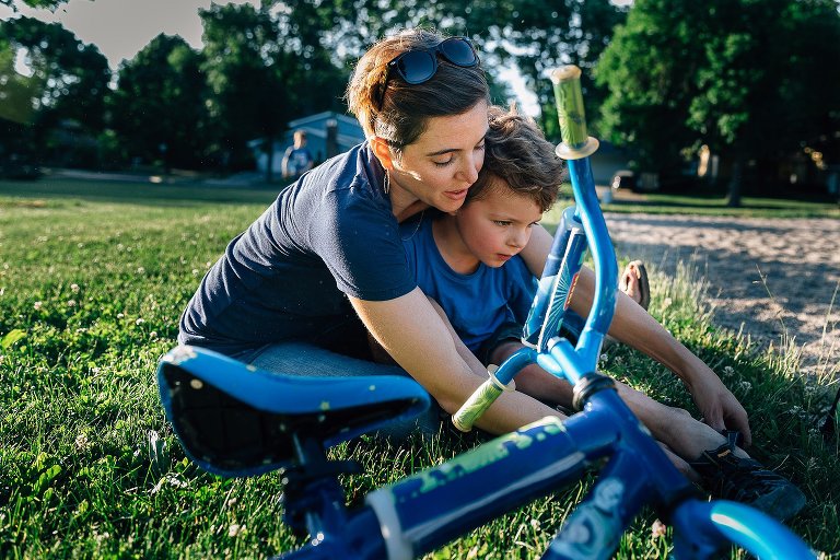 Mother helps son get his shoes on at the park. His bike is in the foreground. 