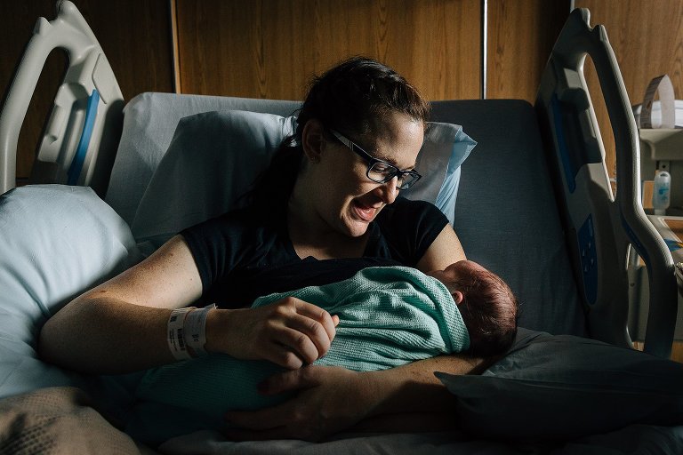 Color. Mother lies in a hospital bed holding newborn child and making eye contact with child. Soft, low light and lots of shadows in the frame. 