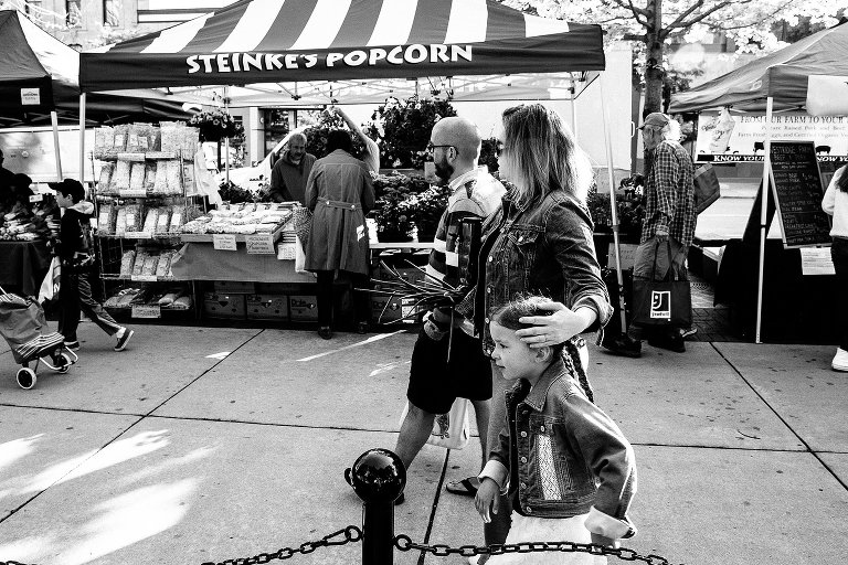 Black and white. Mother gently holds her daughter's head as they walk through a crowded farmers' market. 