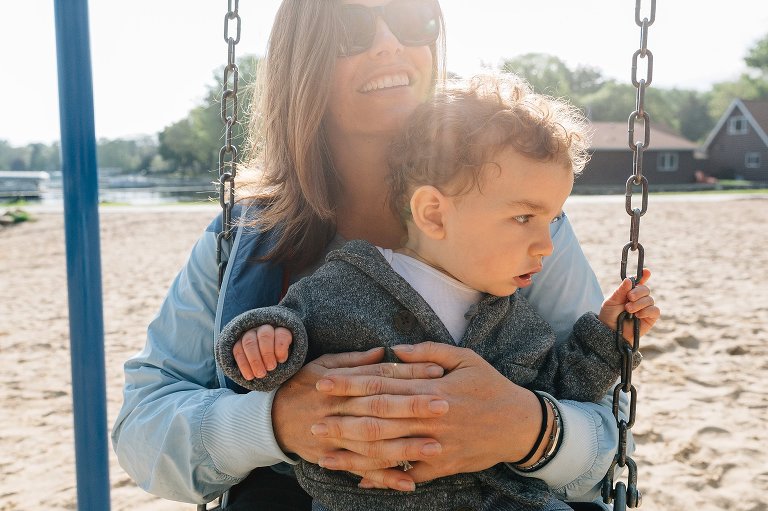 Mother holds toddler son on her lap on the swing. Sun flare coming from top of the frame. 