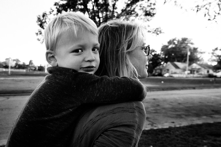 Boy makes eye contact with camera and smiles as he gets a piggy back ride from his mother. 