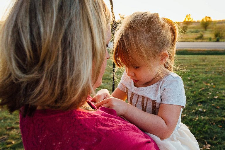 Daughter examines her mother's necklace as they watch the sunset over a field. 