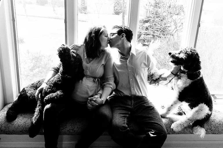 Expectant mother and father kiss in sunny window. Two dogs. 