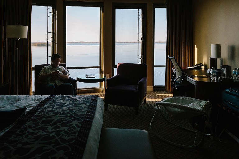 Newborn baby sleeps in dad's arms. They are in a large hotel room on a lake. 
