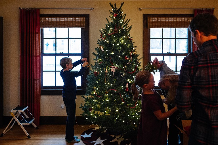 Family decorates Christmas tree in living room, mom hangs mistletoe over daughter and leans in for a kiss